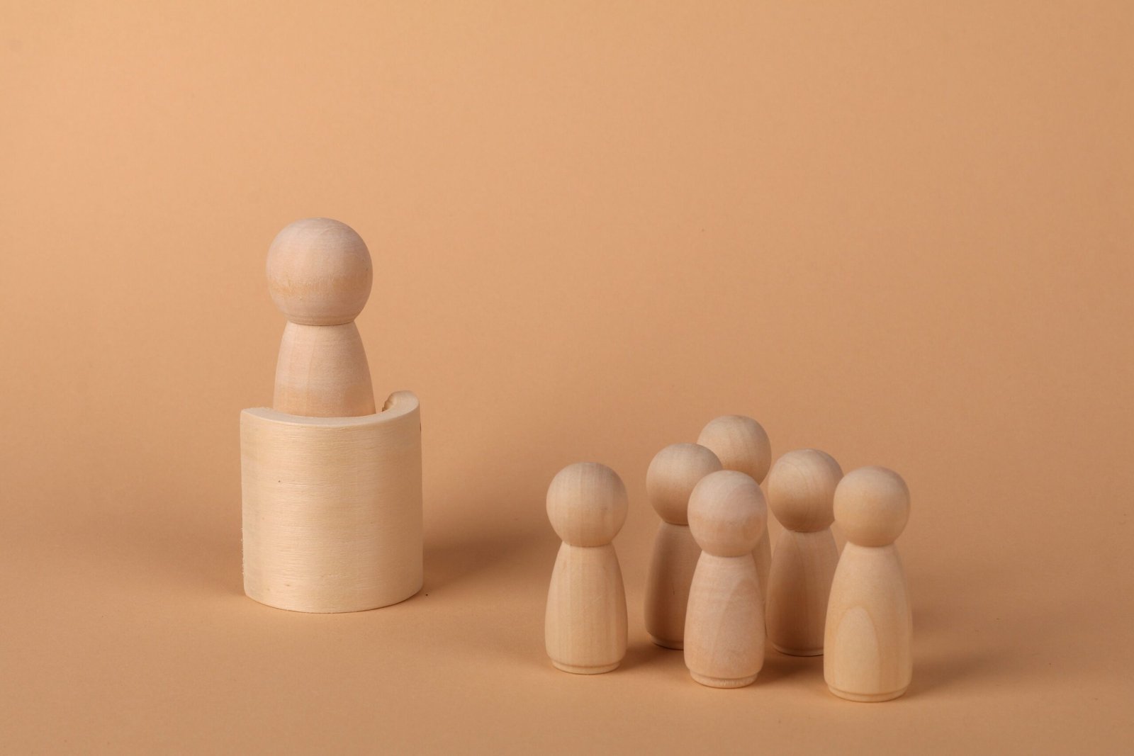 Een houten poppetje in een houder staat apart van een groep, op een abrikooskleurige ondergrond